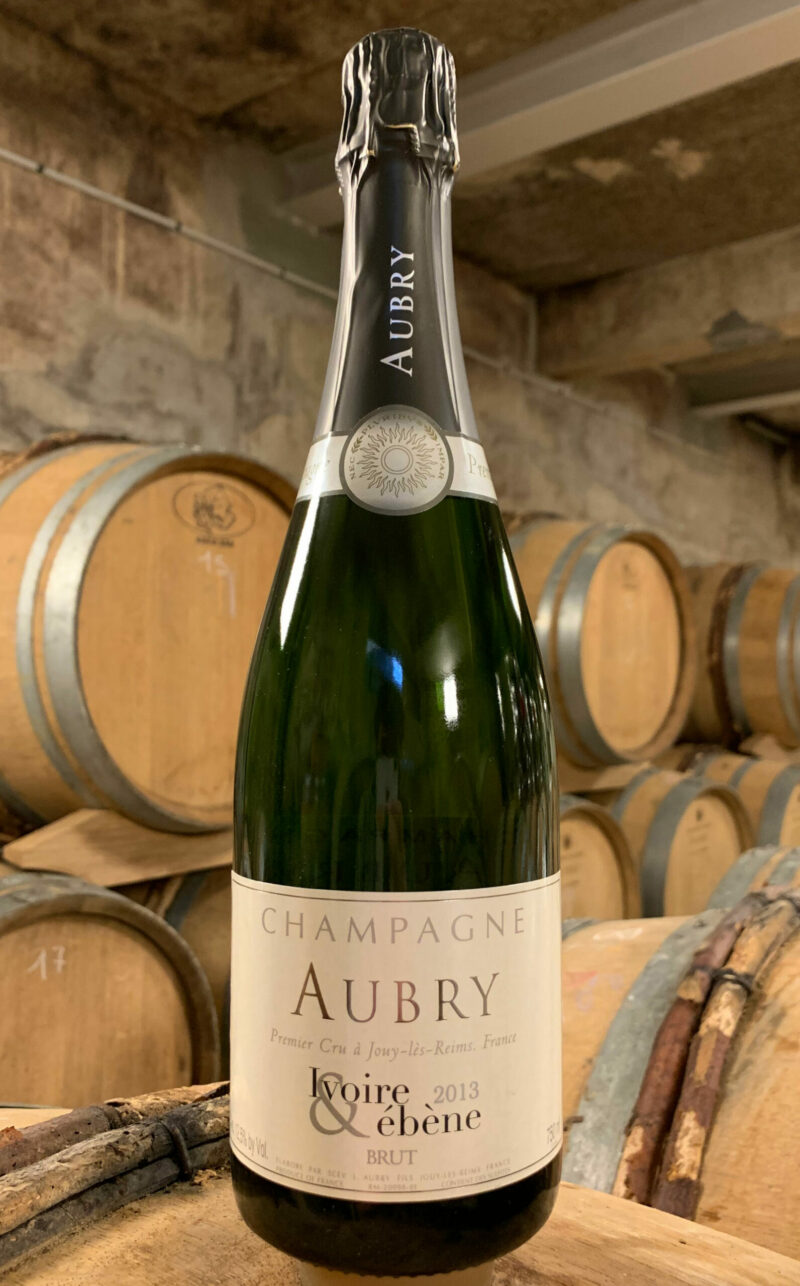 Photo d'une bouteille de Champagne Aubry Ivoire et Ébène prise dans la cave aux côtés des fûts en chêne à Jouy-Les-Reims