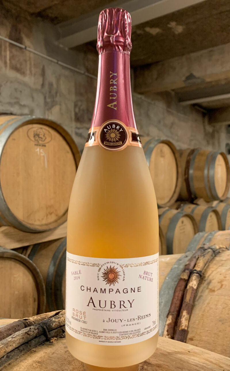 Photo d'une bouteille de Champagne Aubry Sablé Rosé prise dans la cave aux côtés des fûts en chêne à Jouy-Les-Reims